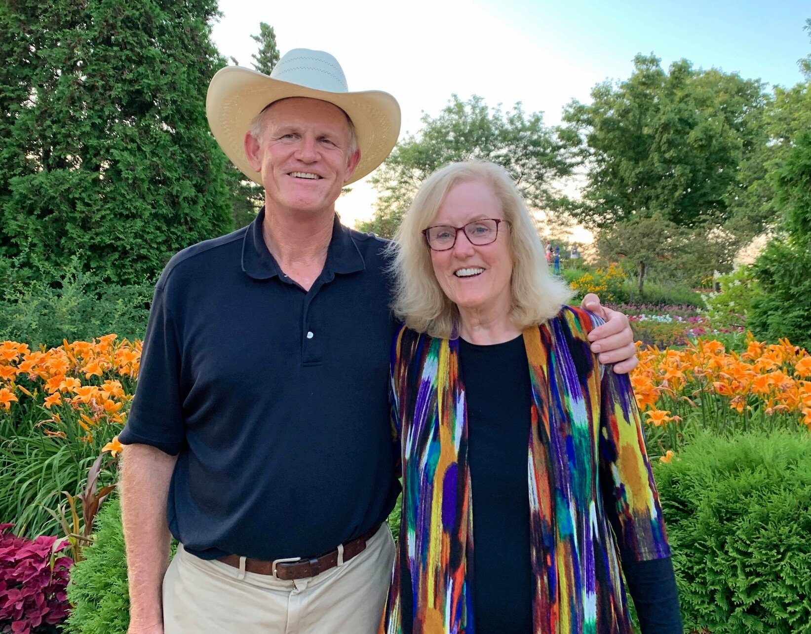 A man wearing a cowboy hat and a woman in a colorful cardigan smile together in a vibrant garden filled with blooming orange lilies and greenery.