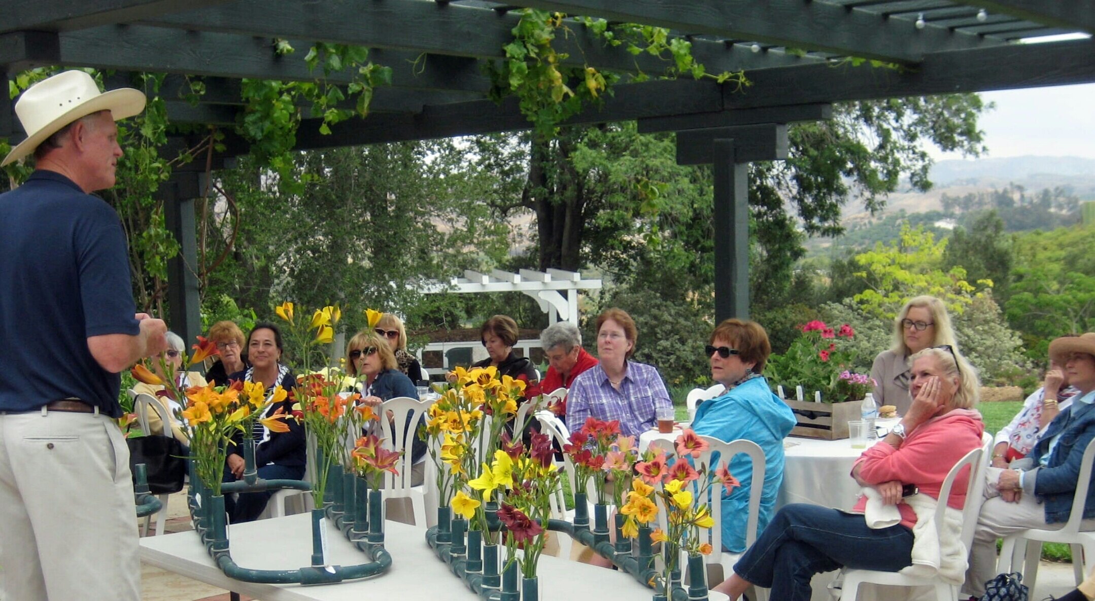 A group of women attentively listening to a speaker during a floral workshop, with colorful flowers displayed on tables under a pergola in a garden setting.